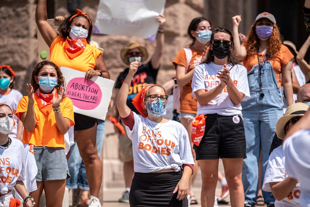 pro-choice protesters in austin yelling and raising their firsts while wearing orange and white shirts that say 
