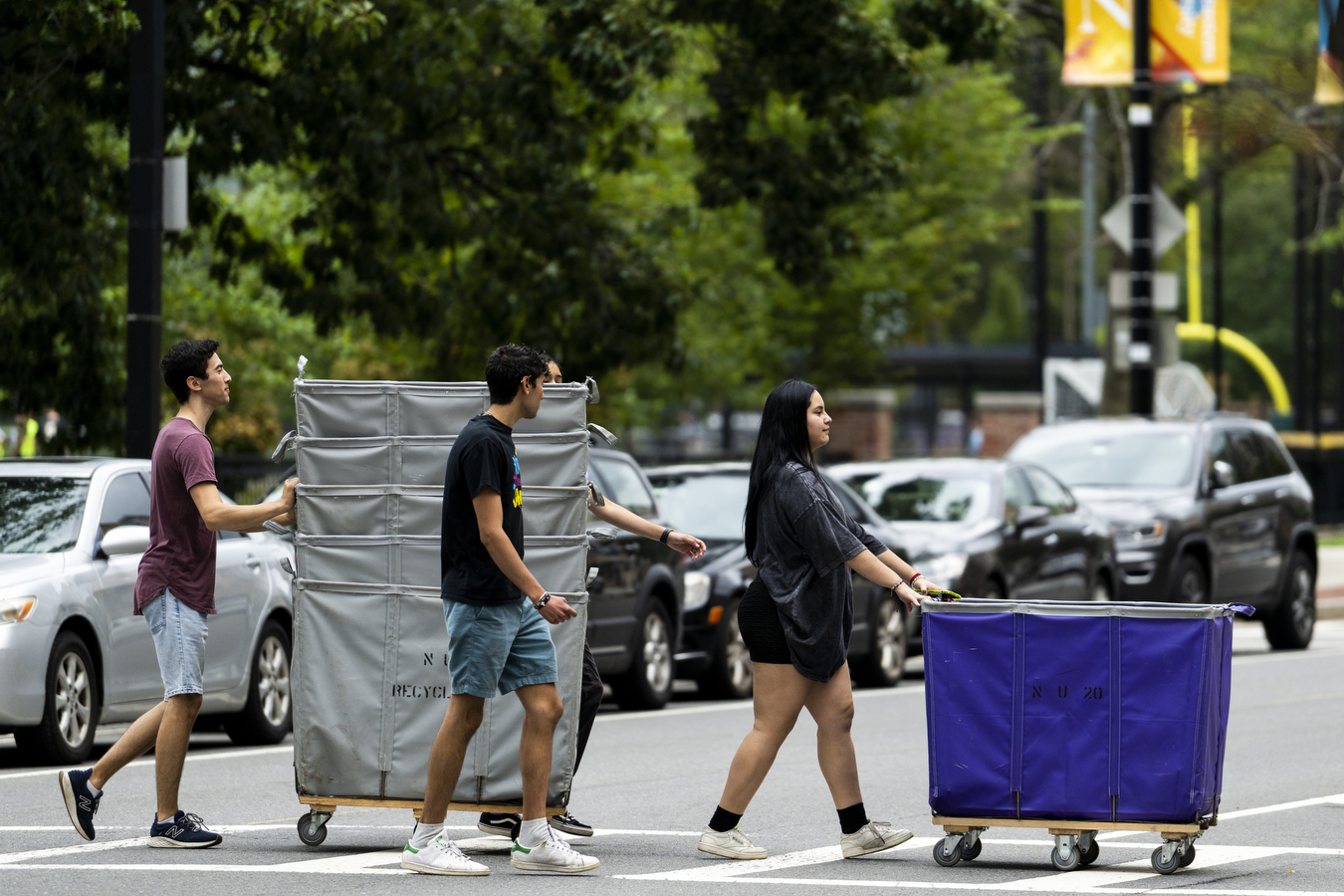 Three people cross the street in a straight line. The first person is wheeling an oversized cart. The second person is walking and the third person is wheeling multiple oversize carts stacked on top of one another. 