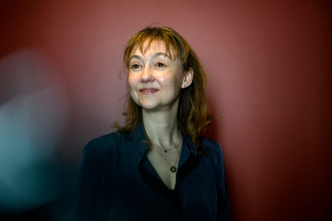 Portrait of Rachel Rodgers wearing a black top and two necklaces in front of a dark red background.