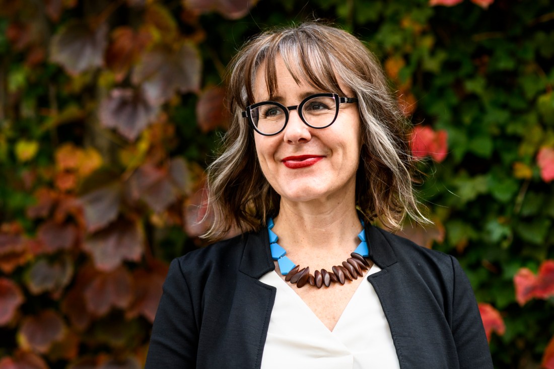A woman with shoulder-length hair, glasses, and a brown and blue beaded necklace smiling for a professional headshot. 