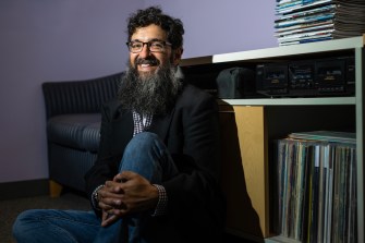 Portrait of Andrew Mall sitting on the ground with one knee propped up. His hands are clasped around that knee.