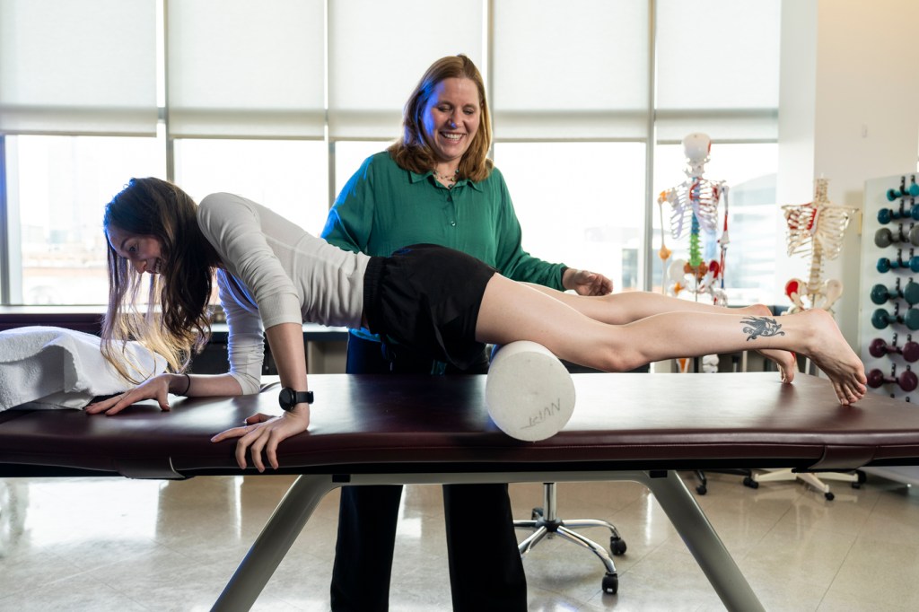 A person stretches on a table with a cylinder foam roller while another stands over them.