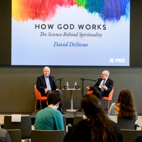 Dave DeSteno and Francis Collins speaking into microphones on stage in front of a screen that says 'How God Works' on it.