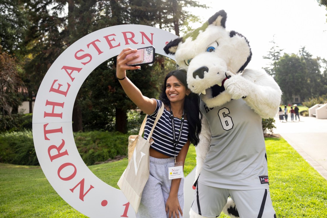 A student poses with Paws the Husky mascot in front of a Northeastern branded decoration on the Oakland campus.