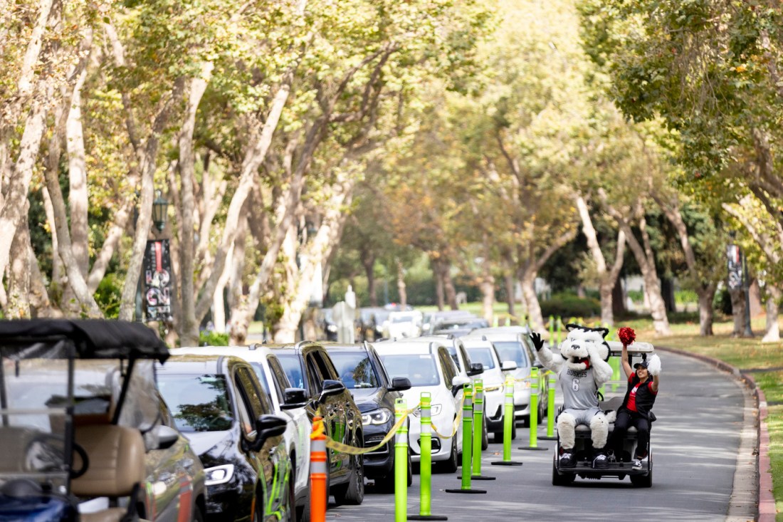 A golf cart with Paws the mascot and his handler with cheer pom poms drives past a line of cars while waving and smiling.