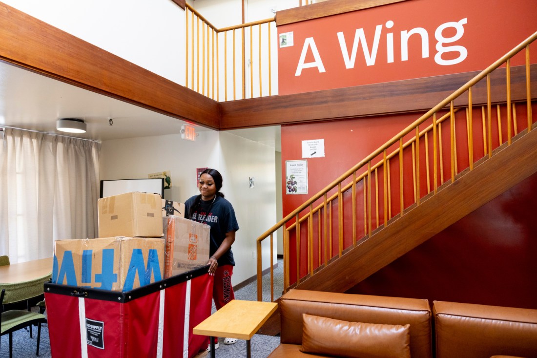 A student pushing a wheeled cart full of boxes through a dormitory common area.