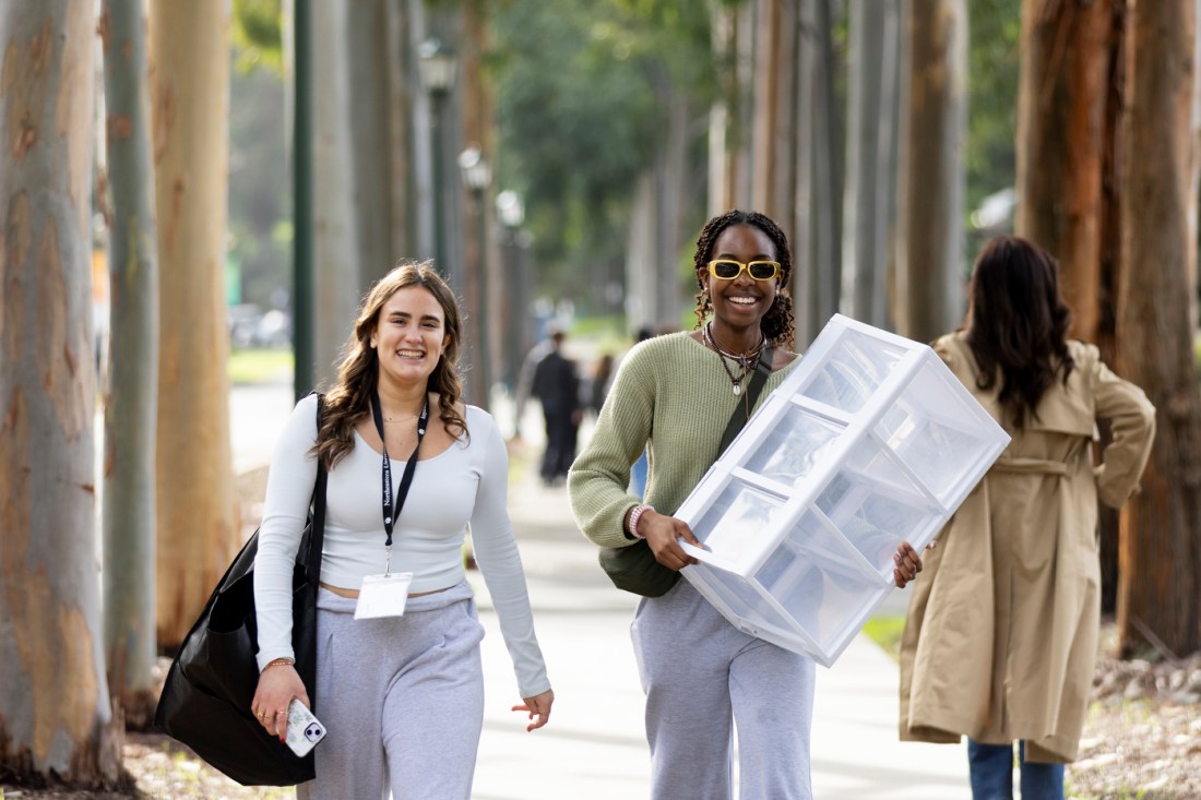 Two students walking towards the camera, one with a large duffel bag over their shoulder and the other holding a set of plastic drawers.