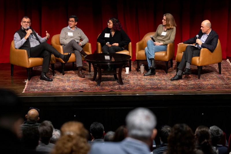 Five people sitting in a semi circle of tan armchairs on a carpeted stage. They are all holding microphones.