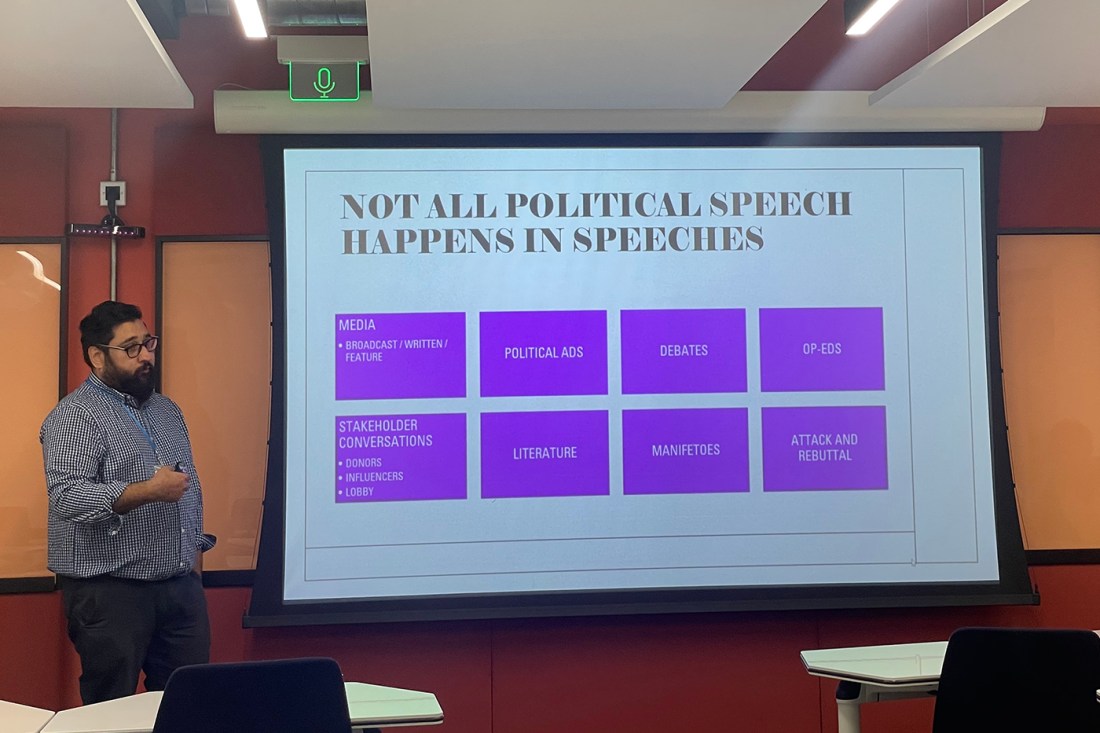 A person with a beard and glasses wearing a blue checkered shirt and black pants standing next to a projector screen. On the screen a slide with a white background is titled 'NOT ALL POLITICAL SPEECH HAPPENS IN SPEECHES' in black text. Underneath, there are eight purple boxes with different speaking points on them, including media, political ads, debates, op eds, stakeholder conversations, literature, manifestos, attack and rebuttal.