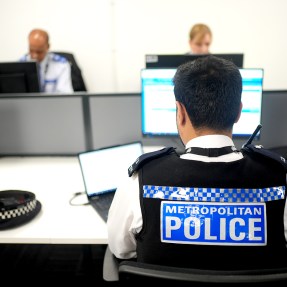 A person wearing a police branded vest sitting at a desk with a laptop and monitor in front of them. On the other side of the desk, two other people sit in front of computers.