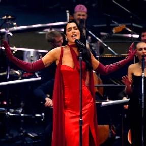 Dua Lipa wearing a red dress while singing into a microphone at the Royal Albert Hall.