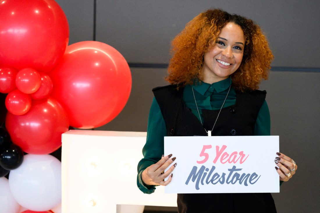 A Northeastern community member posing with a sign that says '5 Year Milestone' next to a bunch of red balloons.