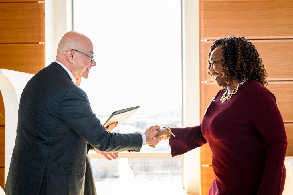 Two people smile and shake hands in a brightly lit indoor space during a formal event, with one holding a document.