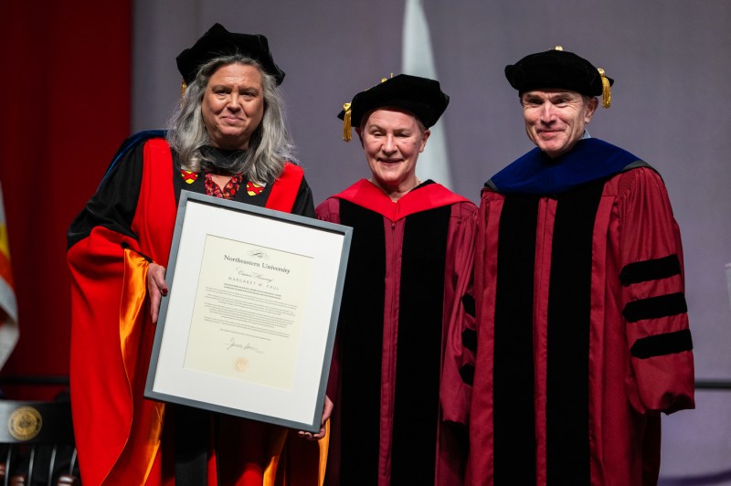 Three people posing on stage with a large frame and an honorary degree inside that frame.