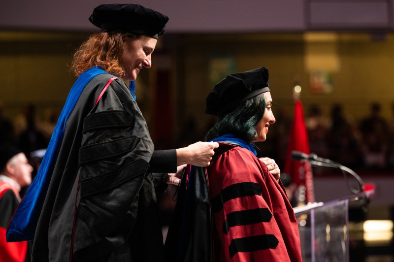A member of the Northeastern community adjusting the regalia of another member on stage at the PhD graduation and hooding.