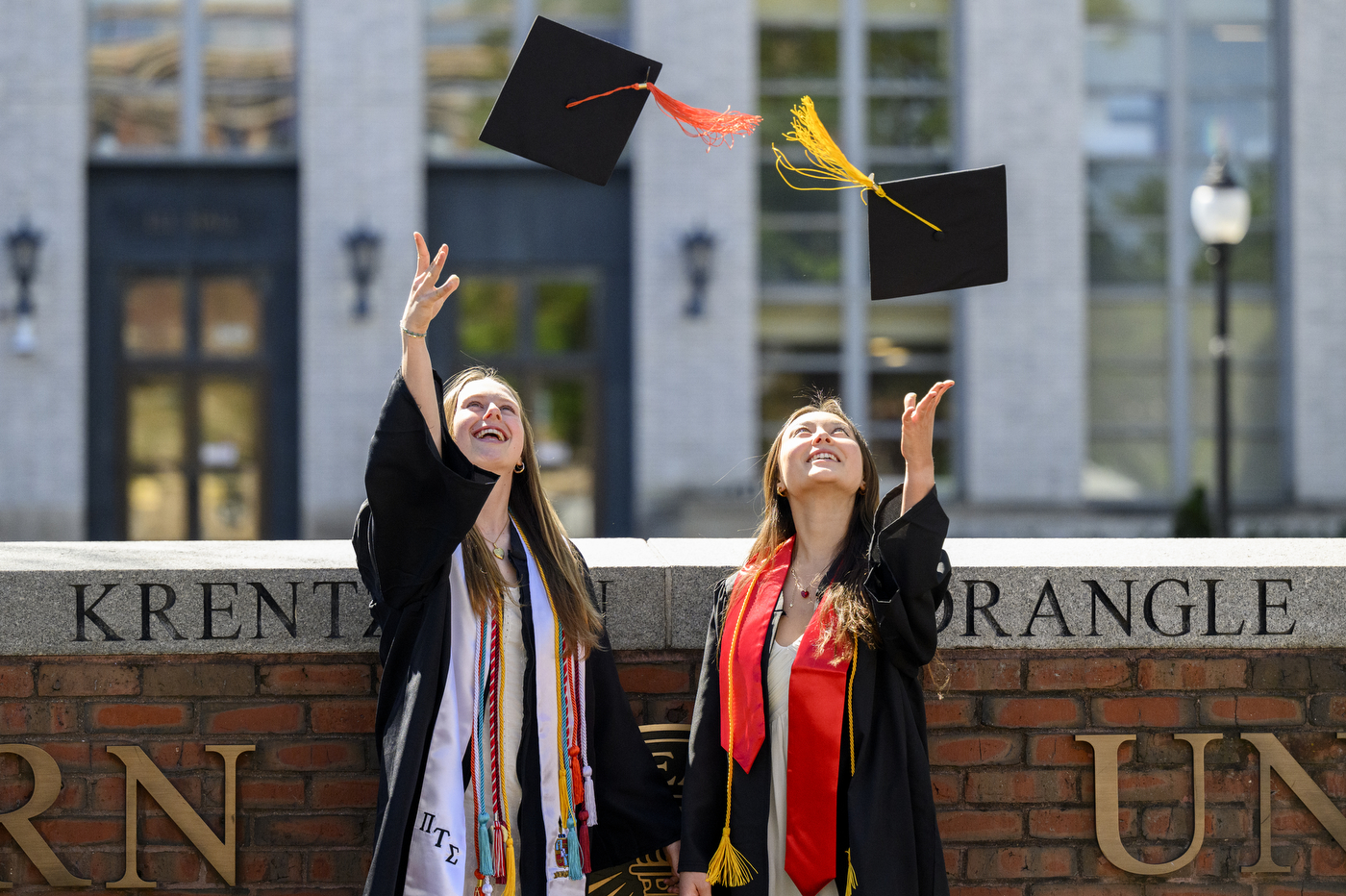 Two people wearing graduation gowns toss their caps into the air while smiling and standing in front of a brick wall on a sunny day.