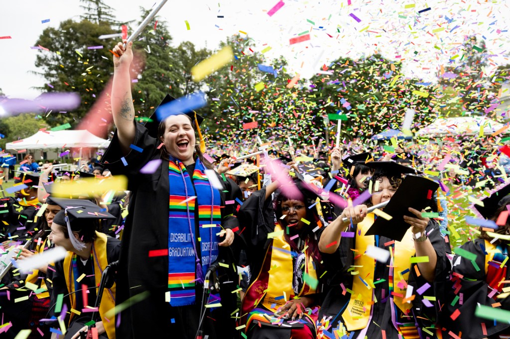 A graduate waving and cheering as confetti falls around them at the Oakland Commencement ceremony.