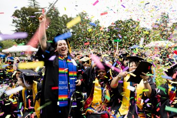 A graduate waving and cheering as confetti falls around them at the Oakland Commencement ceremony.