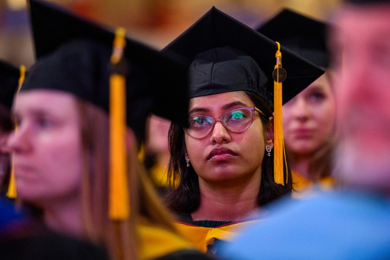 A graduate wearing a cap and gown and glasses listening intently at the CPS Commencement ceremony.