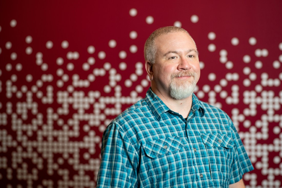 Gary Cantrell, a Northeastern University expert in digital forensics, poses for a portrait in front of a red patterned background.