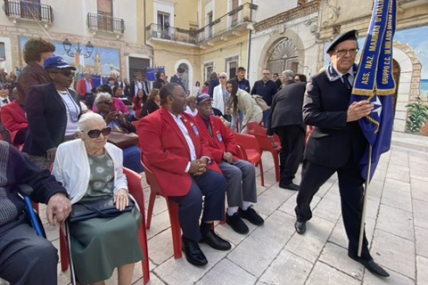 A ceremony in an Italian plaza. Many people are seated and one holds a flag.