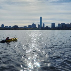 A person kayaks in the Charles River with the Boston skyline visible on the horizon.