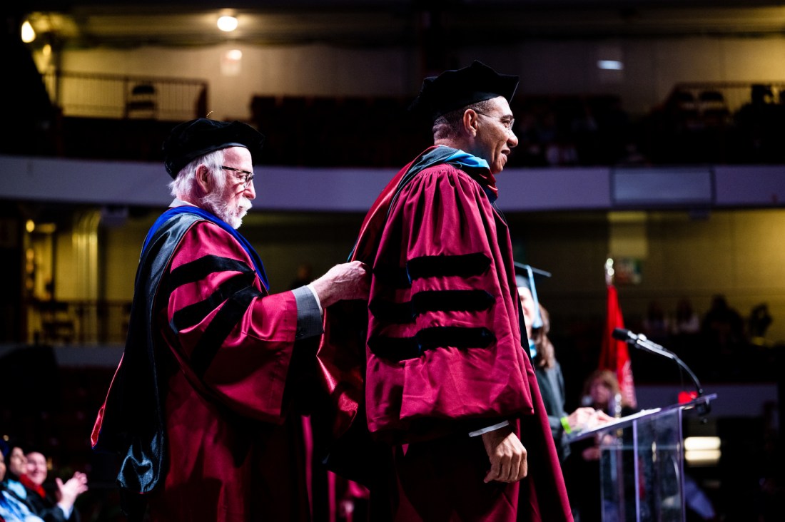 A person adjusting Andrew Michael Holness's commencement robes on stage at commencement.