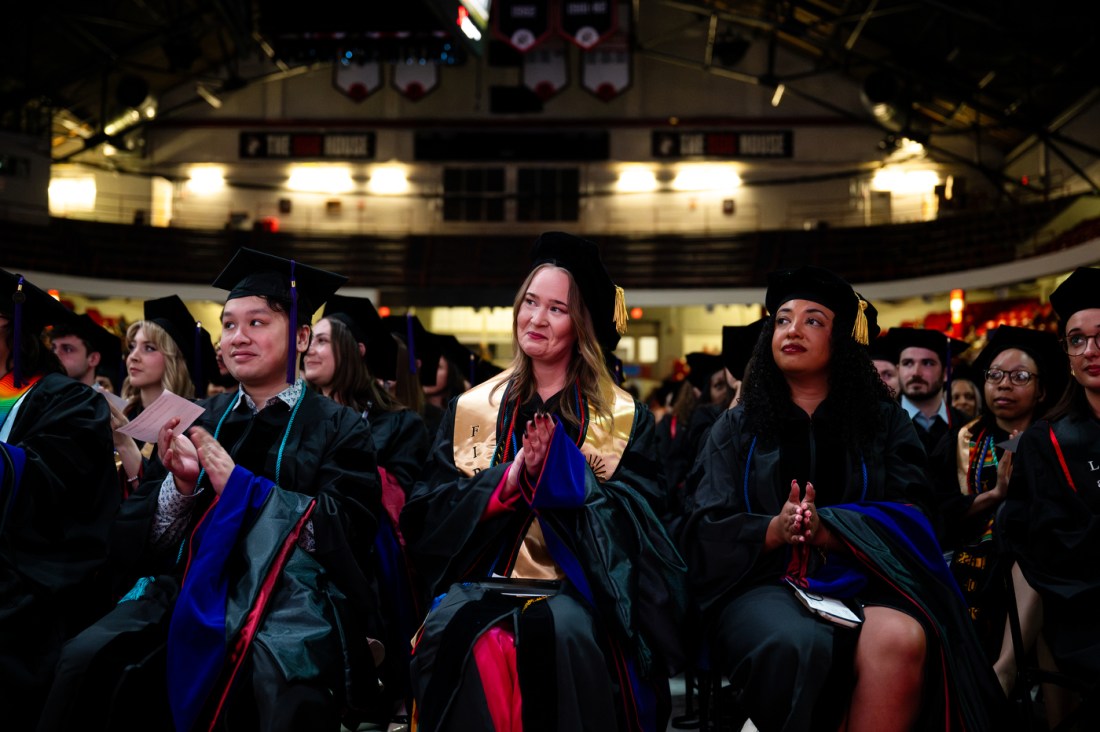 Law school graduates sitting in Matthews Arena wearing their caps and gowns clapping.