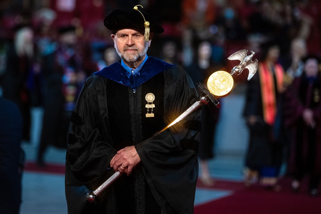 A person in ceremonial academic attire holds a decorative staff while leading a formal procession indoors.