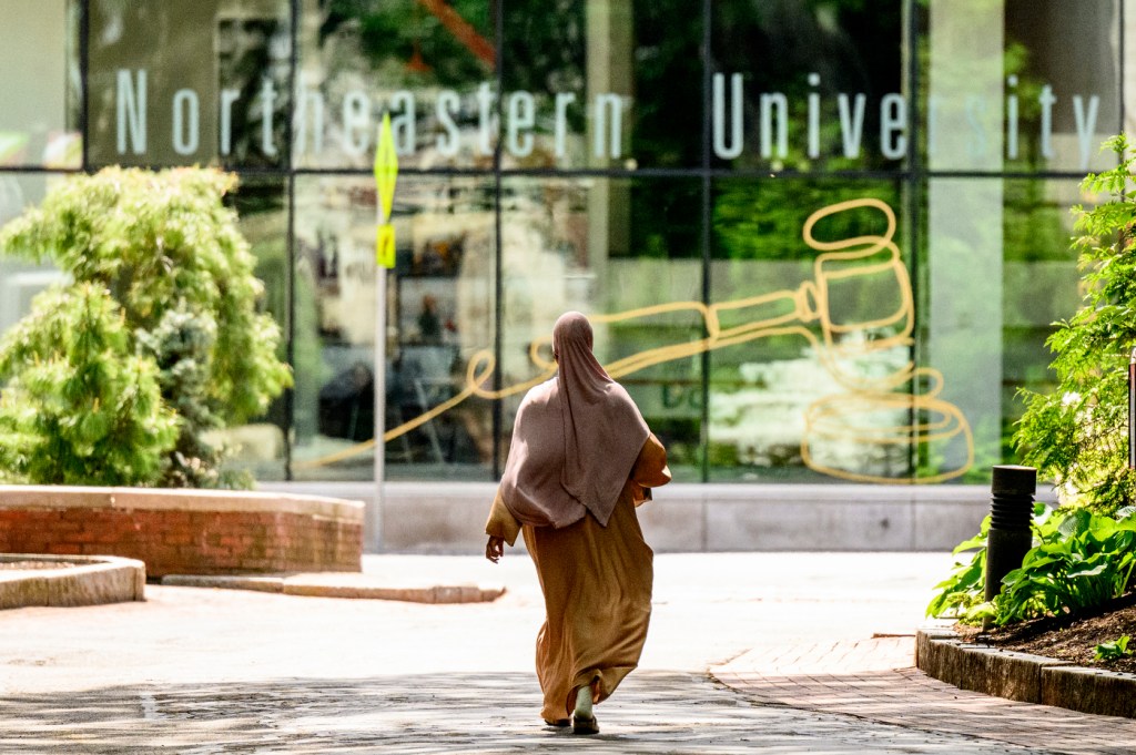 A person walks alone on a shaded pathway toward a building with large windows displaying a university name and a symbolic graphic.