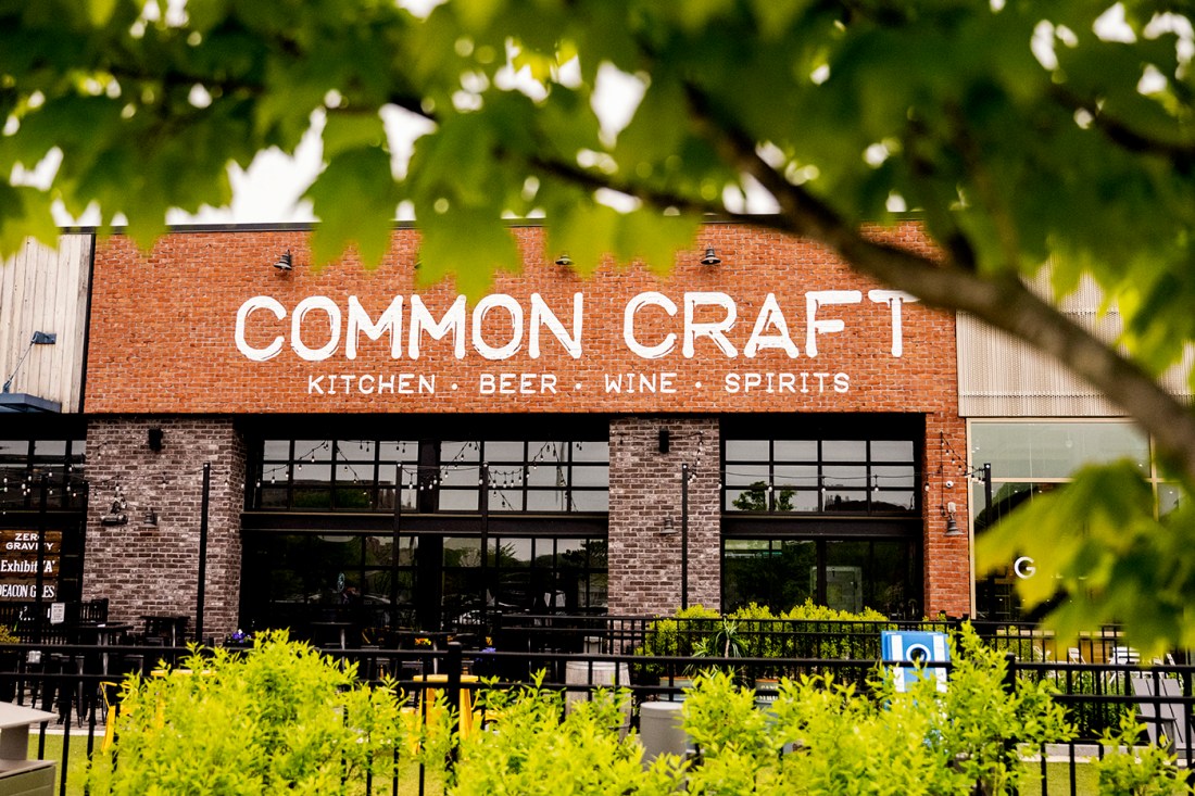 A tree in the foreground of a shot of a brick building that says 'Common Craft' on it in all white, all caps letters.