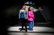 Nita Akoh wearing a pink blazer and blue pants and Jay Nakhai wearing a grey blazer and black pants standing back to back under a spotlight-type light in a darkened room.