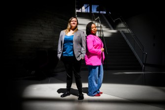 Nita Akoh wearing a pink blazer and blue pants and Jay Nakhai wearing a grey blazer and black pants standing back to back under a spotlight-type light in a darkened room.