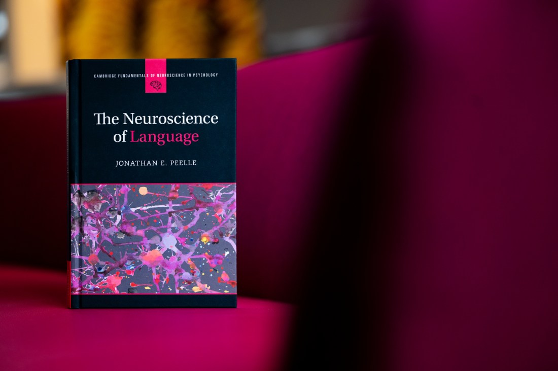 A hardcover book titled "The Neuroscience of Language" by Jonathan E. Peelle is propped upright on a magenta-colored chair. The book cover features a dark background with colorful, abstract neural imagery and is part of the "Cambridge Fundamentals of Neuroscience in Psychology" series.