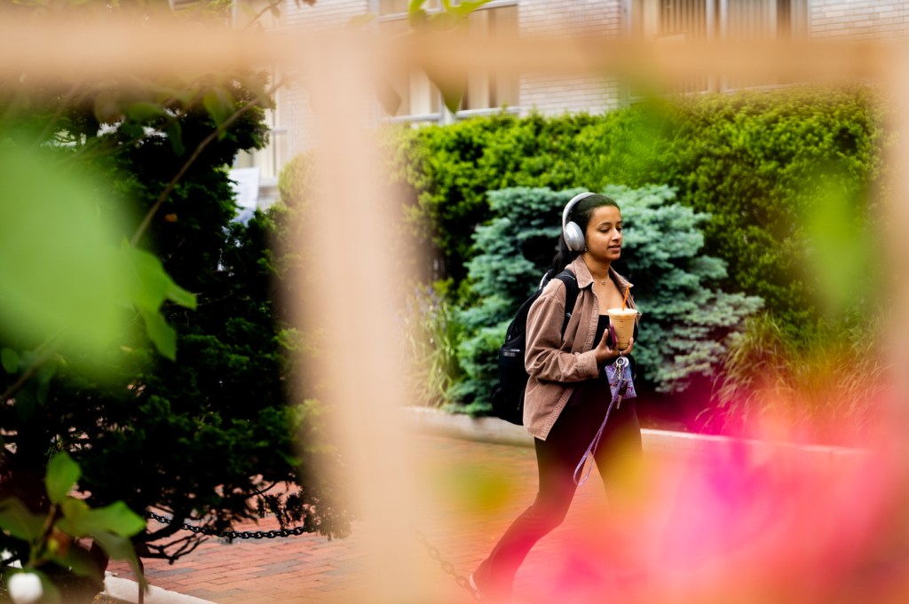 A person walks outdoors with headphones and a drink in hand, framed by colorful flowers and greenery on a brick pathway.