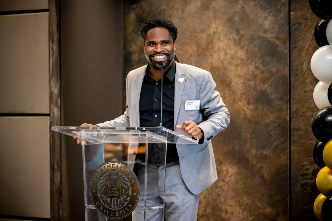 A smiling man in a grey suit and dark shirt stands in front of a textured wall backdrop, with black and white balloons visible in the background during the awards ceremony.