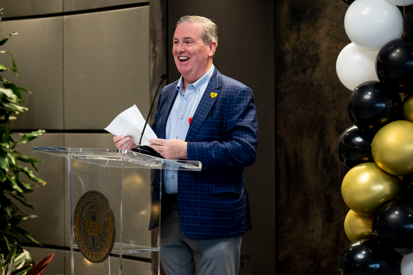 John Tobin stands and smiles in a dark blue suit jacket and light blue shirt at a clear podium, holding papers and speaking at the awards ceremony. Black, white, and gold balloons are visible in the background.