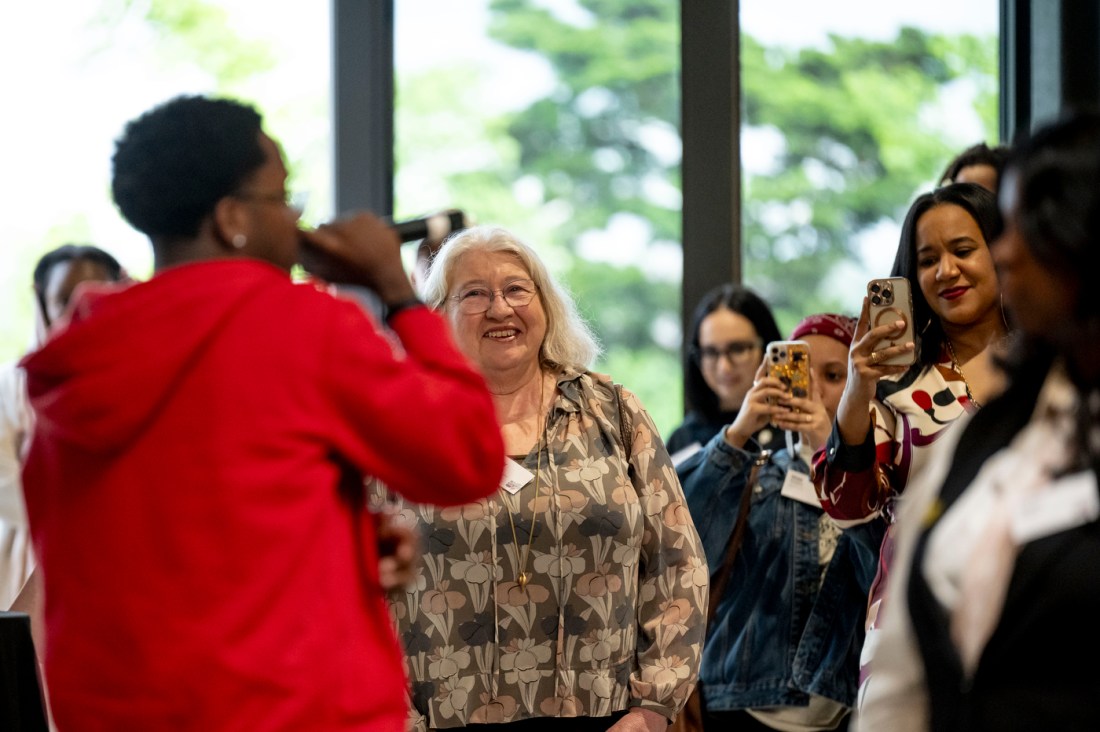 Attendees at the ceremony listen to a person speaking into a microphone, who is wearing a red top and featured prominently in the foreground. Other guests include a woman with white hair smiling and woman with dark hair taking a video of the speaker on her phone.