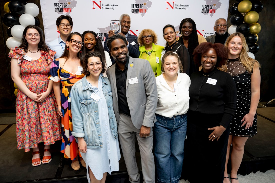 A large group photo featuring approximately 15-20 diverse attendees of various ages posed together in front of a step-and-repeat backdrop with "HEART OF COMMUNITY" and Northeastern University branding. The group includes men and women in business and business-casual attire, with black, white, and gold balloons visible in the background.