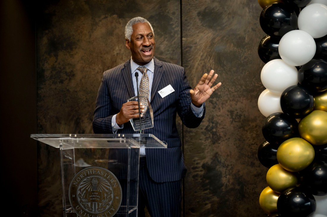 A man wearing a dark suit stands mid speech at a podium. He is in front of a textured wall backdrop with black, white, and gold balloons arranged nearby.