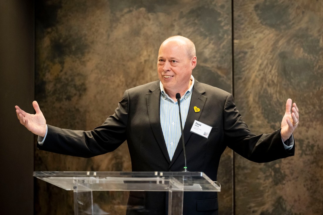 A man wearing a dark suit and light shirt stands before a podium. He is speaking with his hands spread out on either side of him in a gesture. Behind him is a textured wall backdrop.