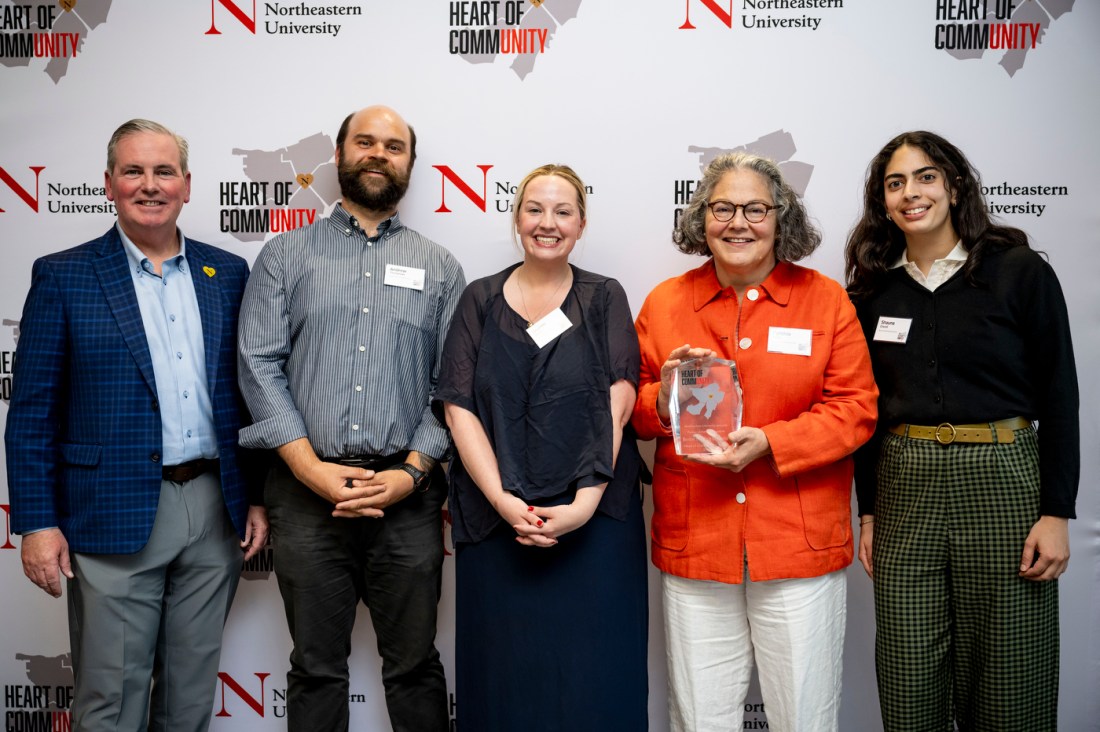 A group photo of five people standing together in front of the branded backdrop. One of the people is holding a crystal award trophy. They are all wearing name tags and smiling at the camera.