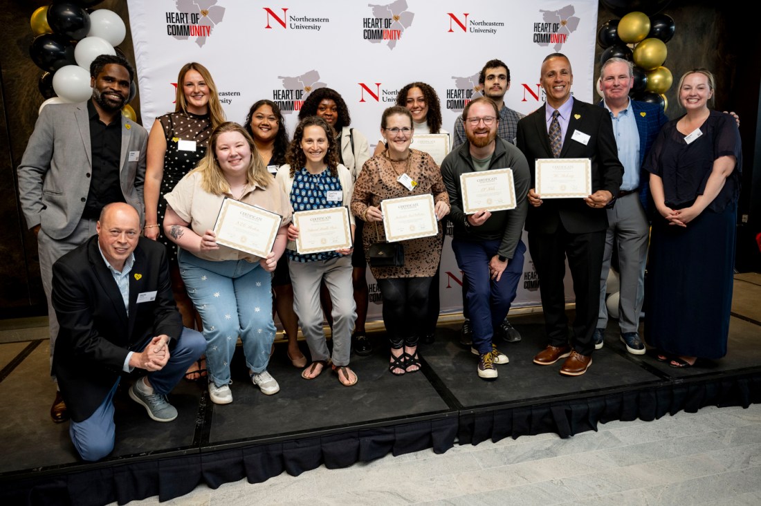 A group photo of approximately 15 award recipients and ceremony organizers pose together in front of a step-and-repeat backdrop with "HEART OF COMMUNITY" and Northeastern branding on it. Several people in the front row are holding certificates or awards. There are black, white, and gold balloons visible in the background.

