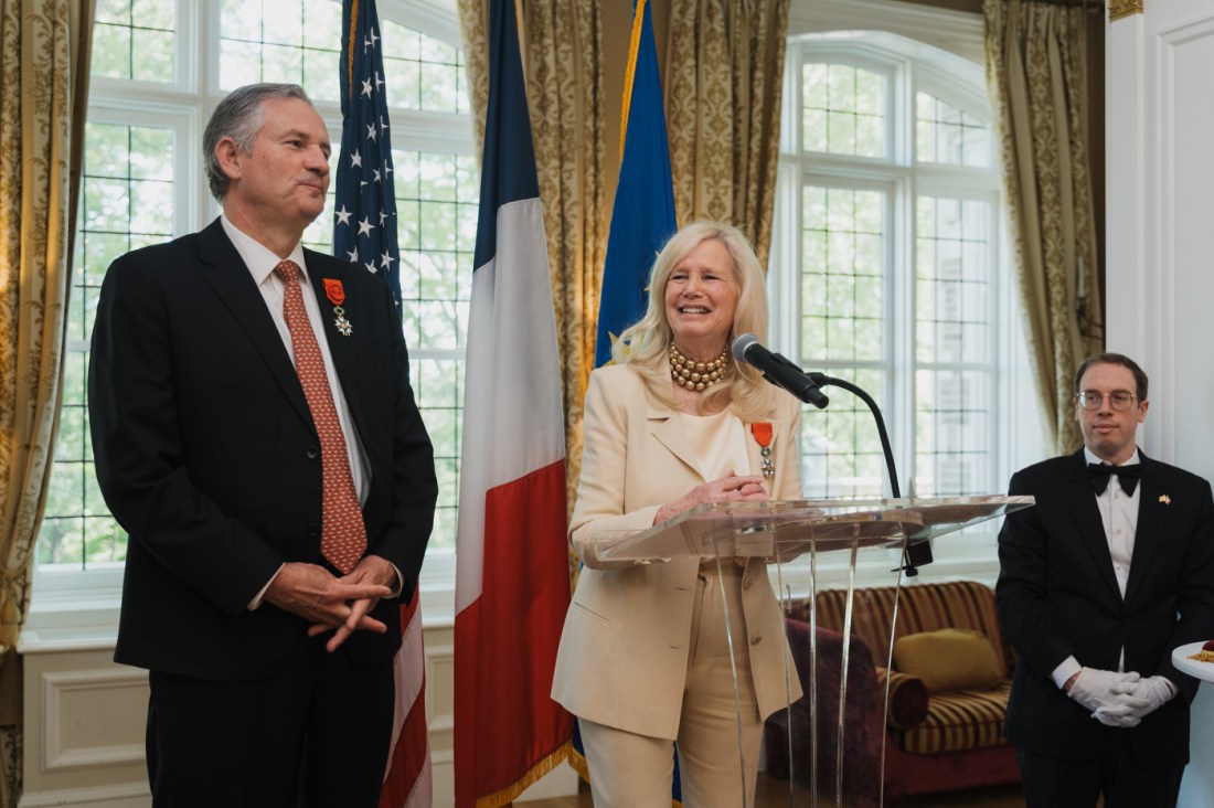Susan Blumenthal stands at a podium smiling as French Ambassador Laurent Bili looks on during an award ceremony at the Embassy of France.