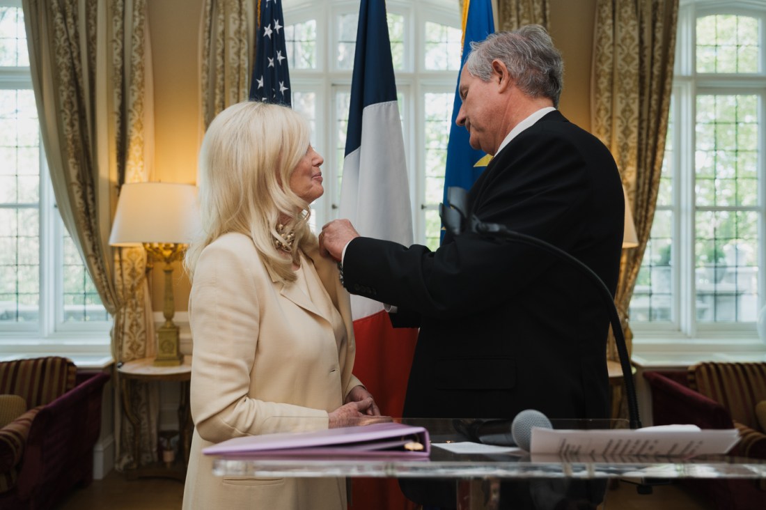 A man presents France’s Legion of Honour medal to Susan Blumenthal during an official pinning ceremony in front of U.S. and French flags.