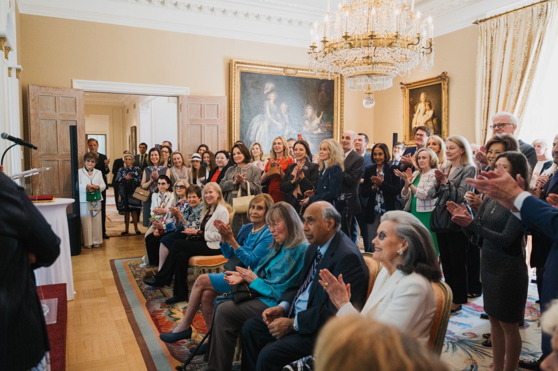 A large group of people, seated and standing, applauds during a formal ceremony in an ornate room with chandeliers and paintings.