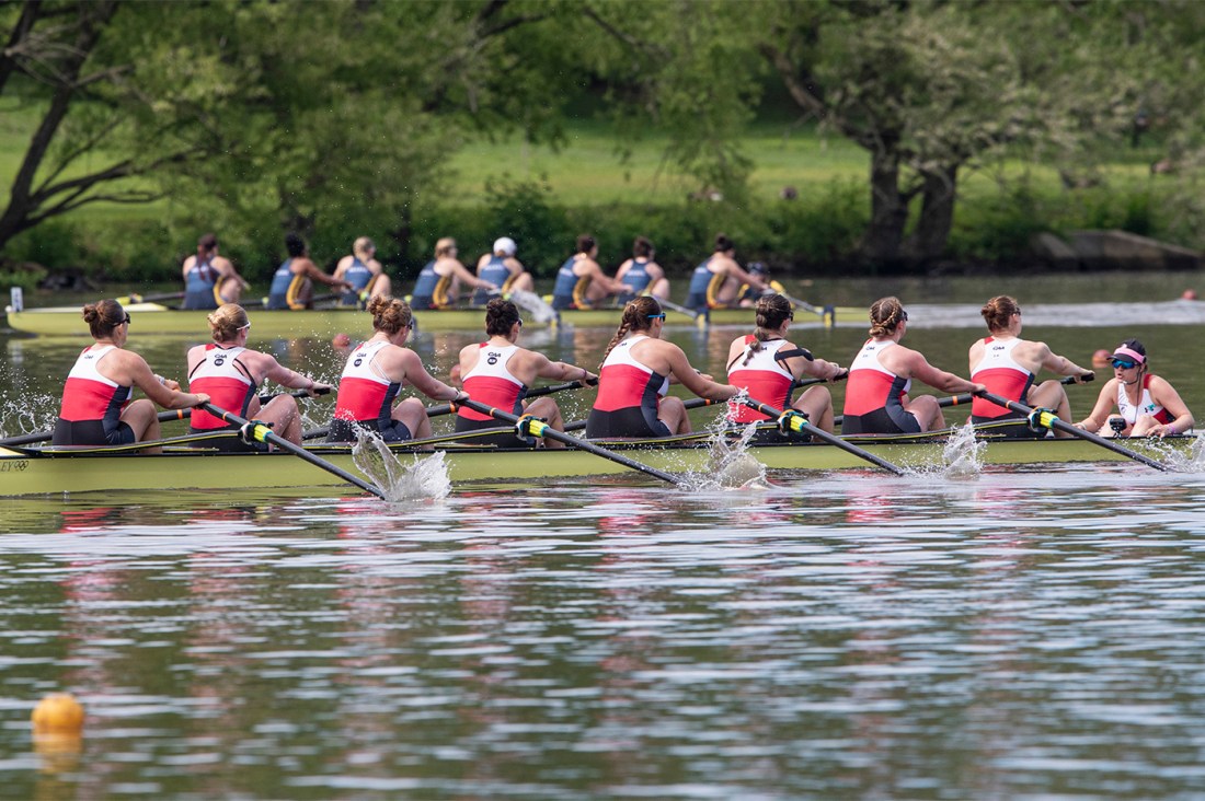 A women's rowing team wearing white and red uniforms with black shorts rows in unison in a yellow boat on a river during a race. Trees and spectators are visible in the background.
