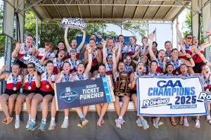 Members of the womens row team sit on a stage cheering after winning the CAA finals. They are holding signs that say 'CAA Champions 2025' and 'CAA Ticket Punched' on them.