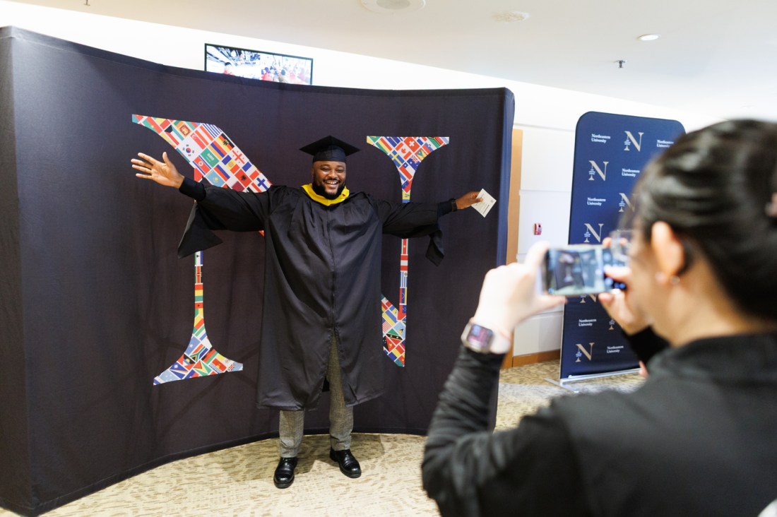 A graduate poses with arms outstretched in front of a decorated photo backdrop as another person takes their picture during Northeastern University Vancouver Campus's Convocation 2025.