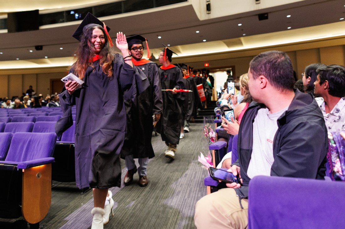 Graduates in black caps and gowns wave enthusiastically while walking through an auditorium filled with purple-velvet seating and audience members.